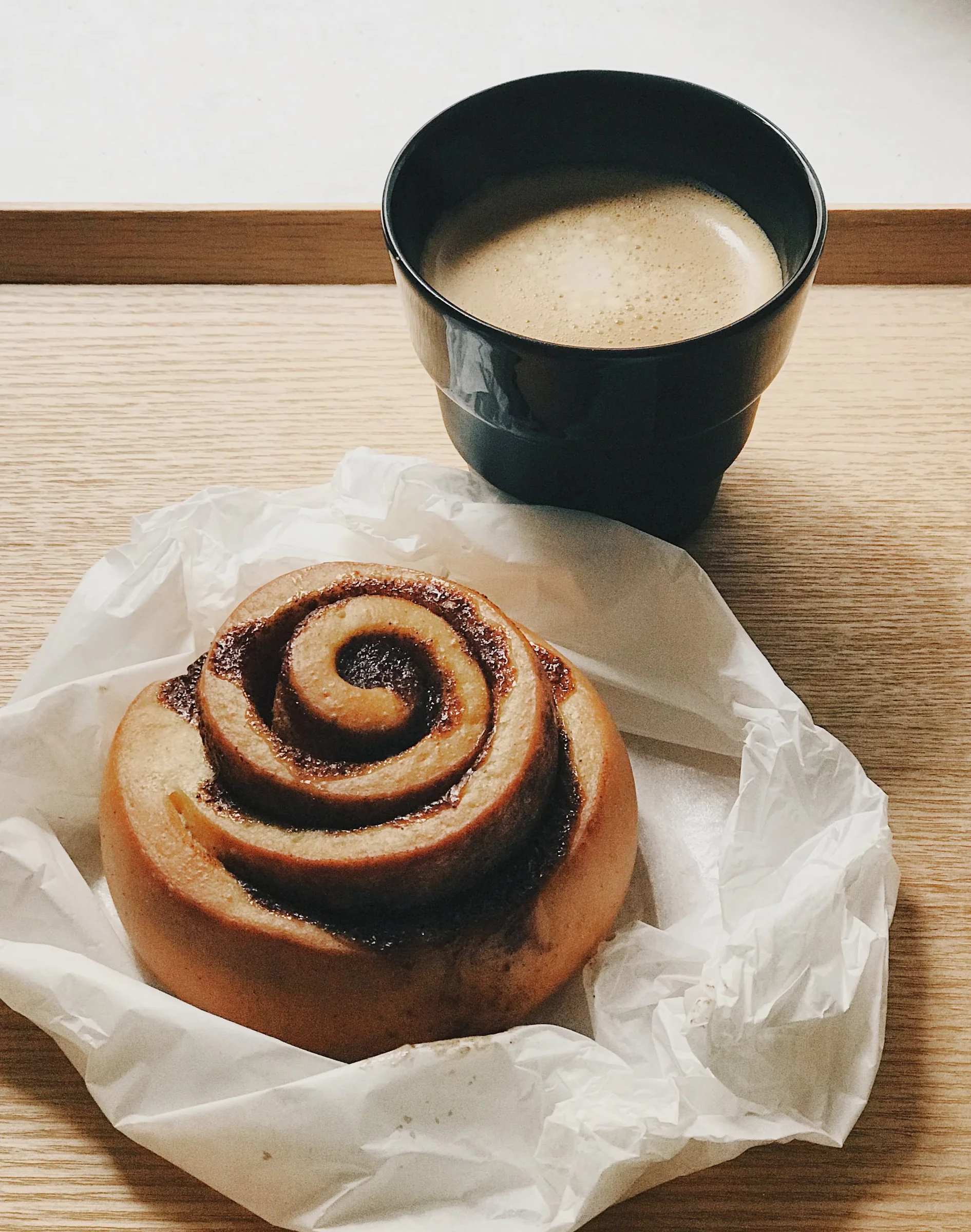A cinnamon roll on paper beside a cup of coffee
