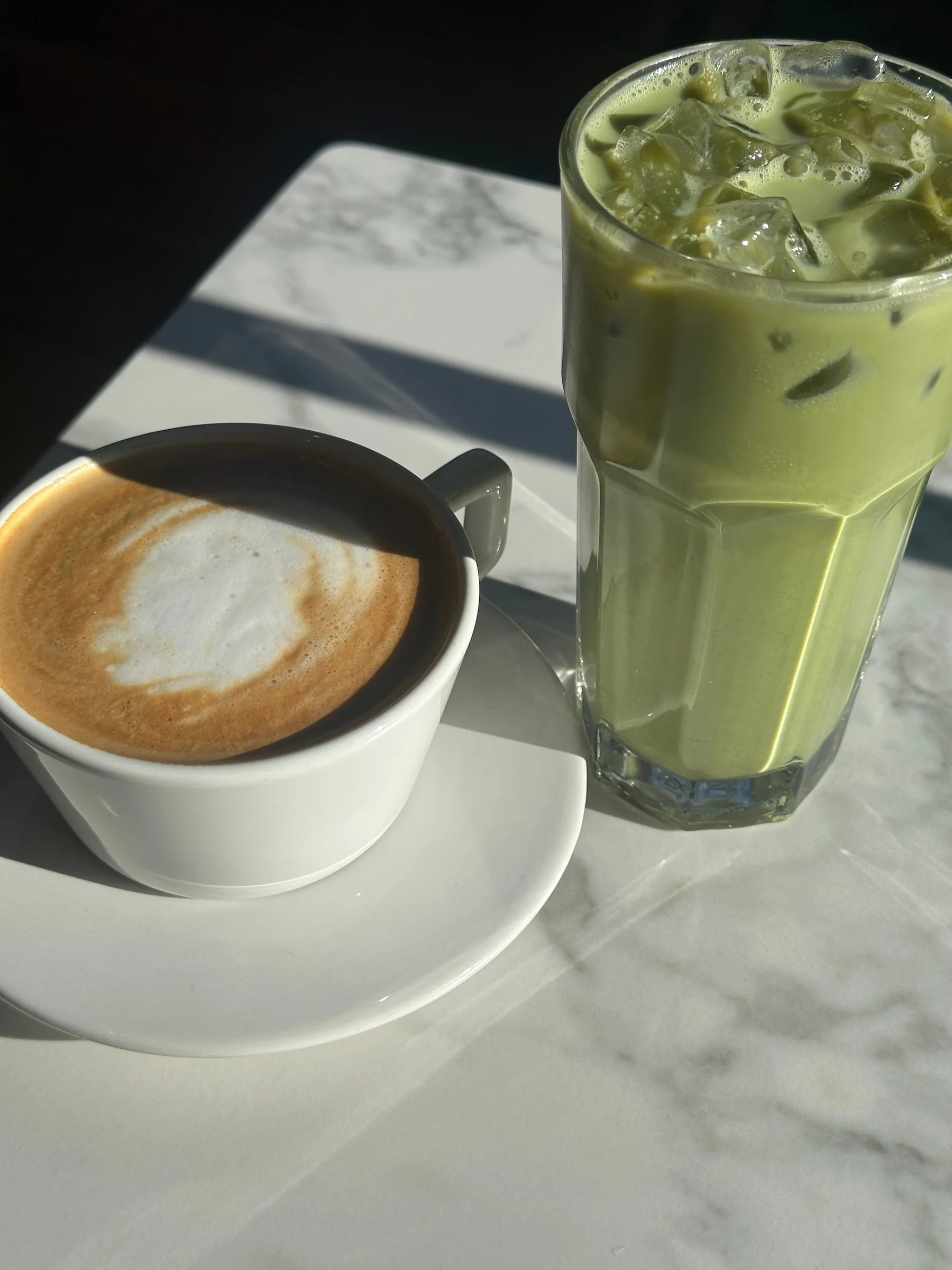 An iced matcha latte beside a cappuccino on a sunlit marble table