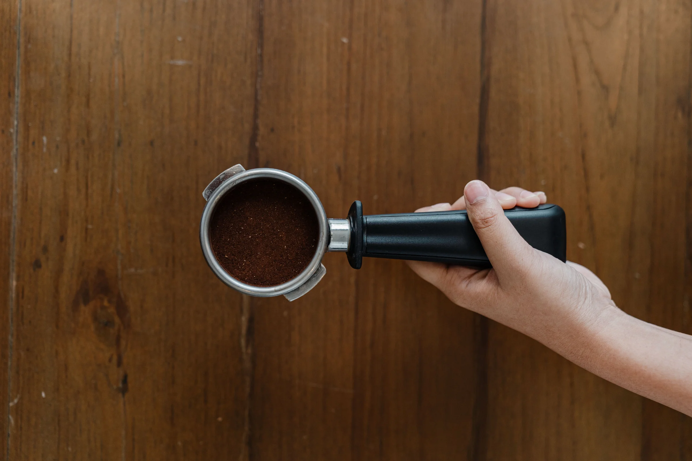 A hand holding a portafilter of freshly ground coffee over a wooden counter